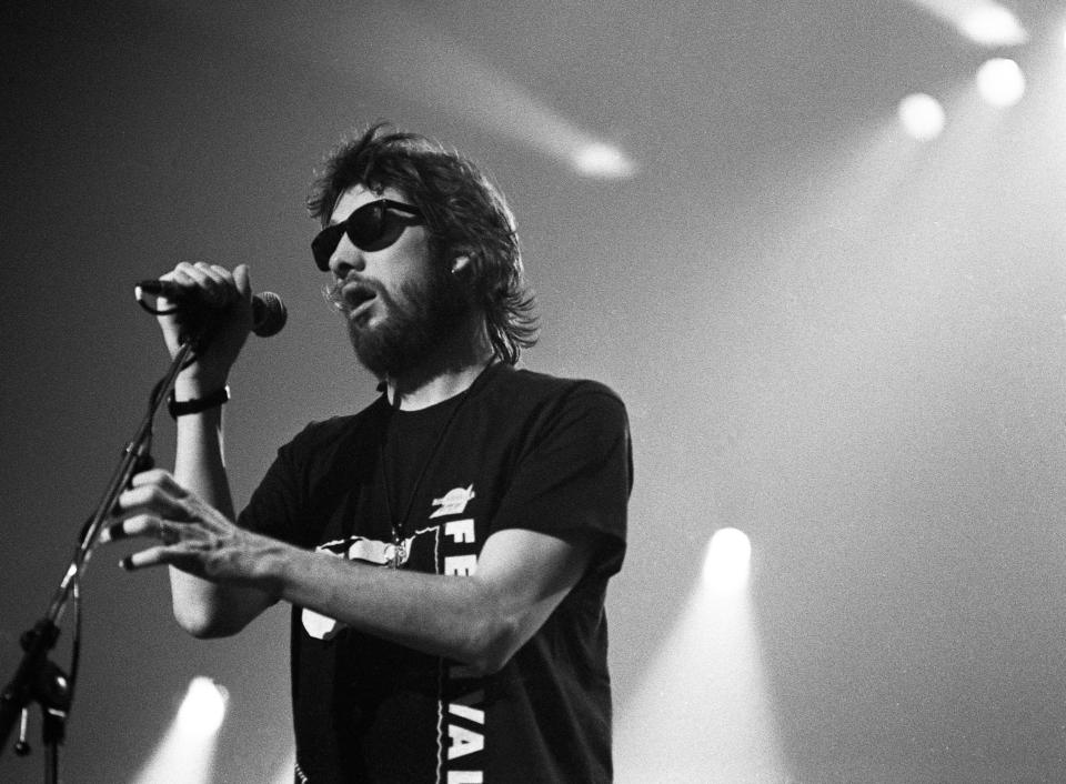 Shane MacGowan in sunglasses holding a microphone in stage