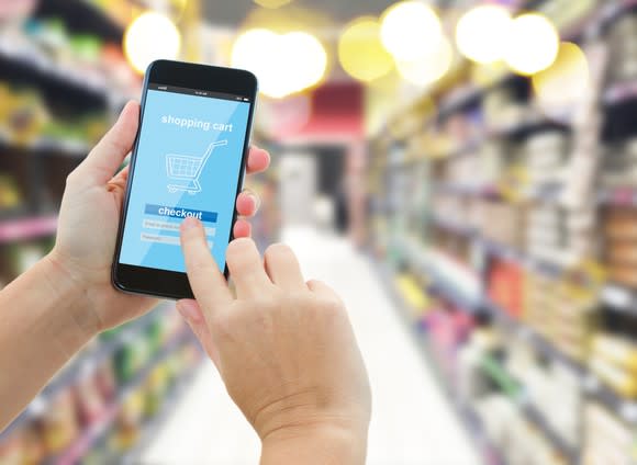 A hand taps a shopping cart icon on a cell phone of a person standing in a grocery aisle.