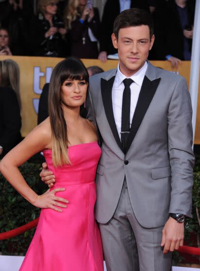 Lea Michele is wearing a pink strapless dress and is posing with Cory Monteith who is in a gray suit with black trim