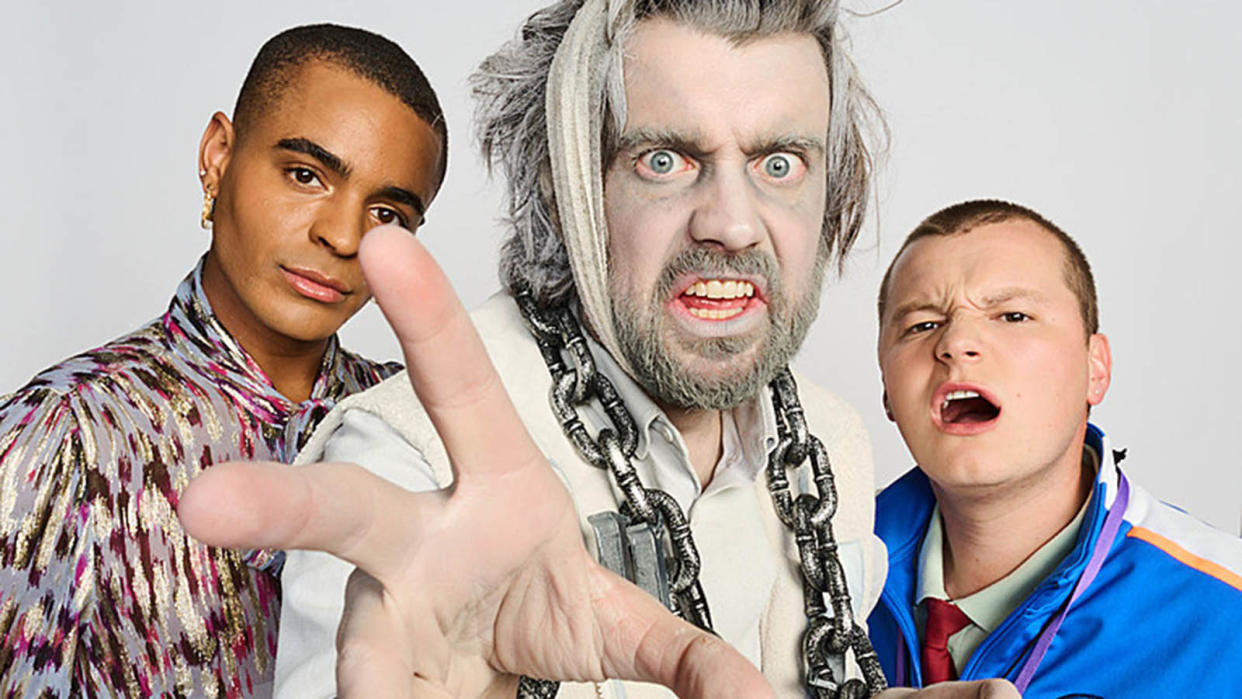  A still from Bad Education: A Christmas Carol showing Stephen (Layton Williams) and Mitchell (Charlie Wernham) standing behind Alfie Wickers (Jack Whitehall). Alfie is wearing grey make-up with his hair and stubble tinted white, and he has large metal chains hanging around his neck. 