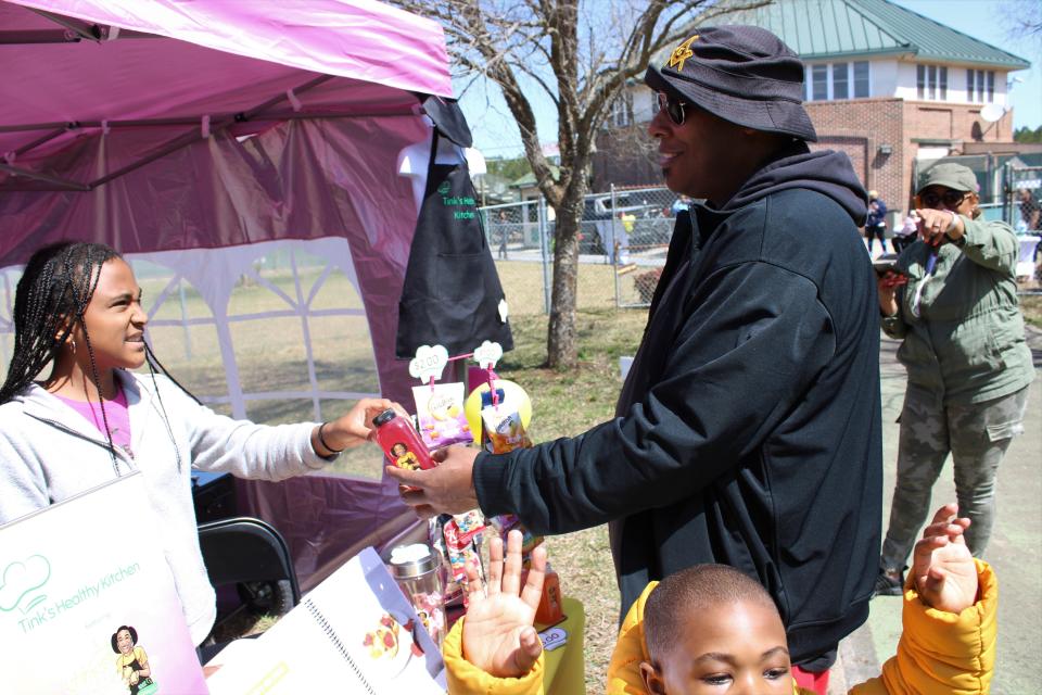 Daquan Reinhardt purchases a drink from Amanni at her Tink's Healthy Kitchen booth during Donamatrix Day at Petersburg Sports Complex on April 2, 2022. Reinhardt's four-year-old son Da' Mir is photo bombing.