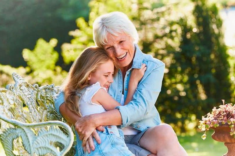 An older woman is holding her granddaughter as they sit on an outdoor bench together, smiling.