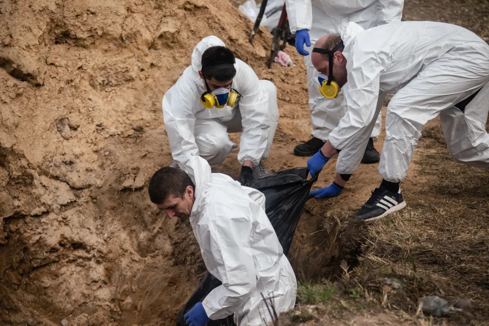 Several men, dressed in white and some wearing masks, pull a body out of a ditch.