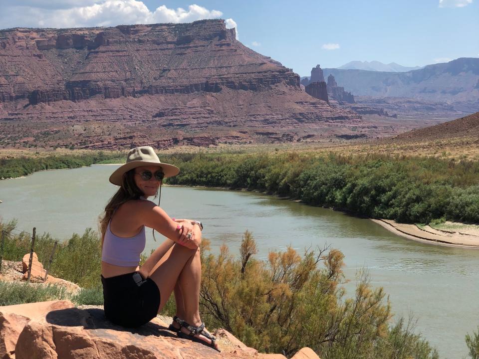 Emily, wearing a purple crop top, black shorts, a beige hat, and sunglasses, sits on a rock looking out at a small body of water and large rock formations.