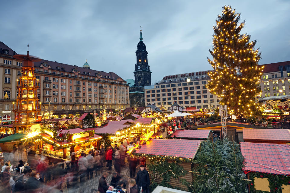 Crowded holiday market with festive lights and a large decorated tree, near a historic tower