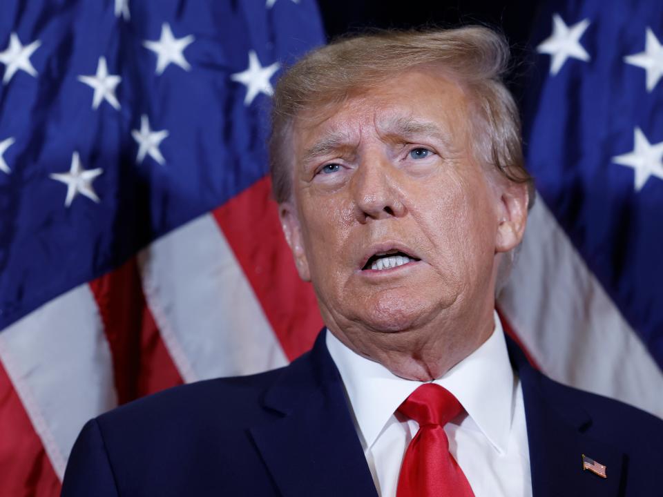 Former President Donald Trump standing in front of an American flag while wearing a suit and tie.