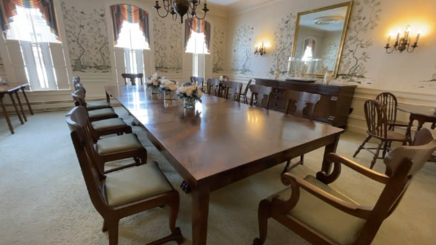 A long mahogany dining table stands surrounded by matching chairs and three vases holding white flowers.