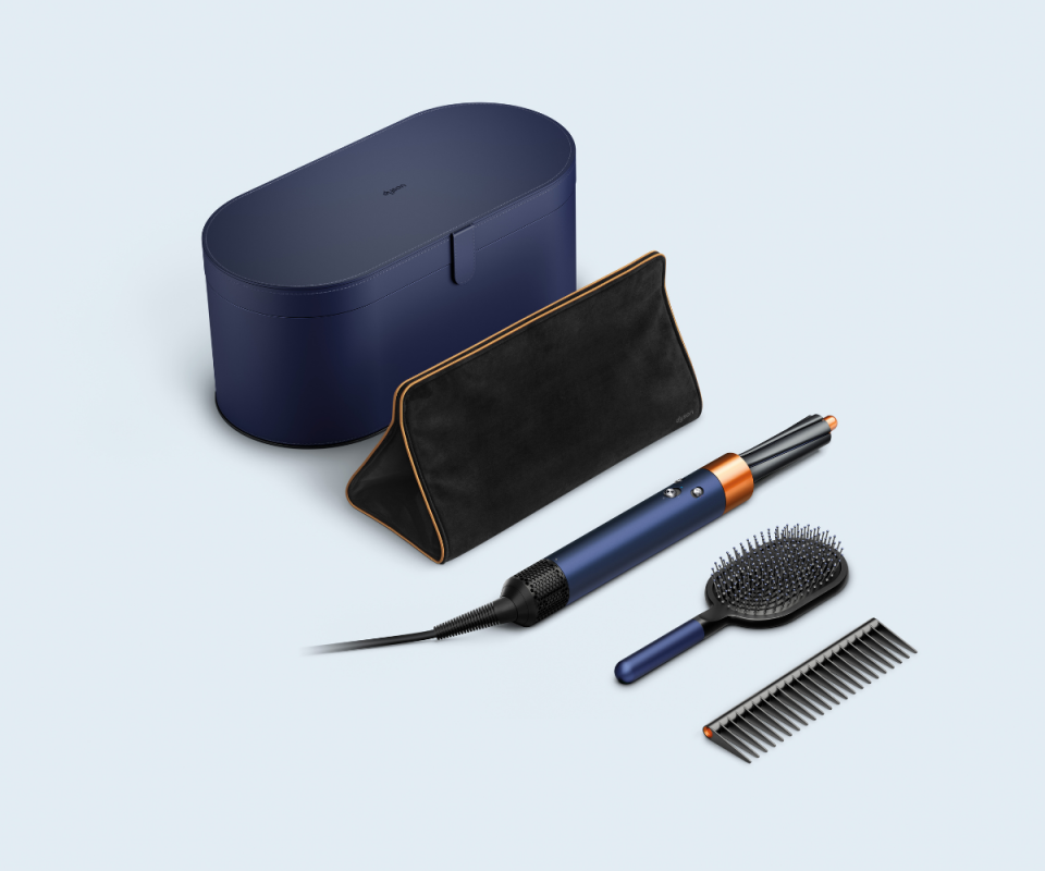 A collection of Dyson's Prussian blue gift box with a black heat mat, airwrap, black brush and comb against a pale blue background.