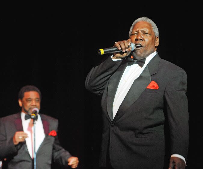 A man with short gray hair wearing a black suit and bowtie and singing into a microphone