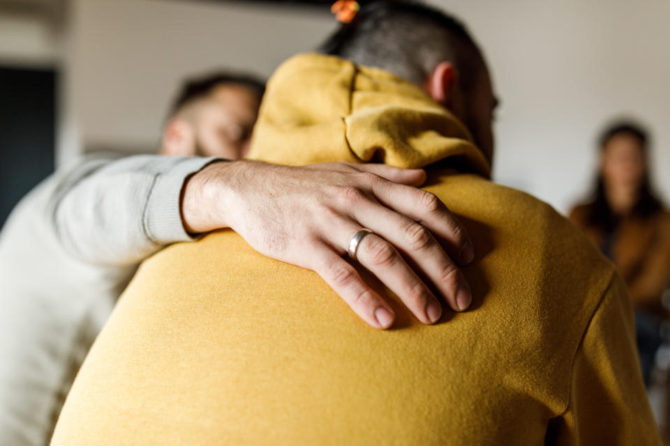 A person wearing a ring gives a comforting pat on the back to someone in a hoodie. Other people are blurred in the background