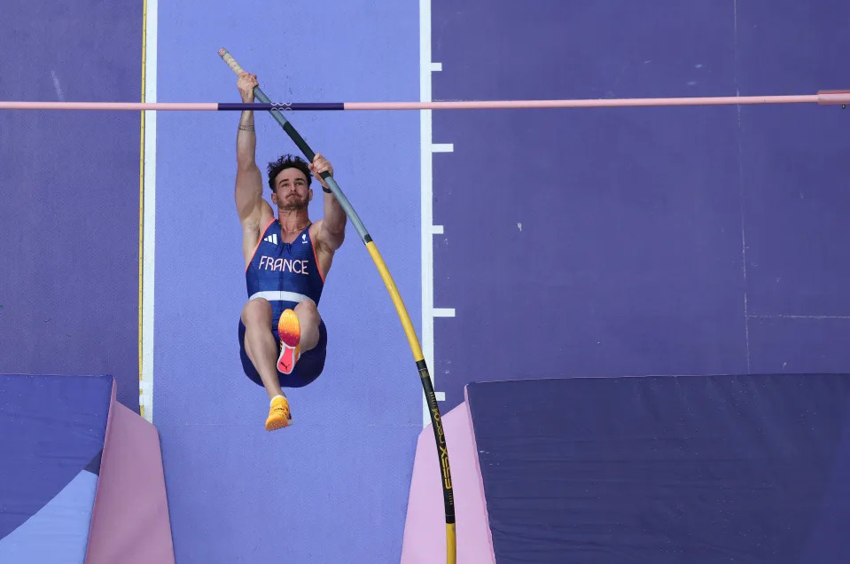 A male athlete in a "France" uniform is pole vaulting, extending his body upwards with the pole in mid-bend during a competition