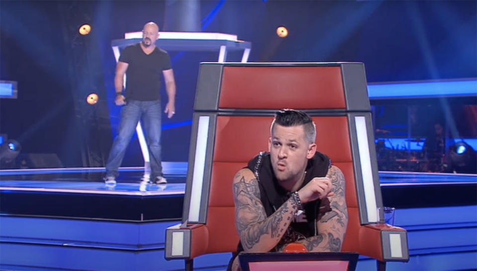 Joel Madden in The Voice red chair with Michael Stangel singing behind him.