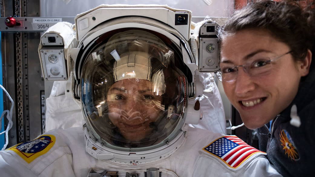  One astronaut in a spacesuit and another unsuited beside her, both smiling, in a selfie picture. 