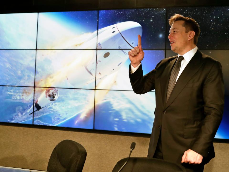 Elon Musk in a black suit, pointing up, in front of a giant screen