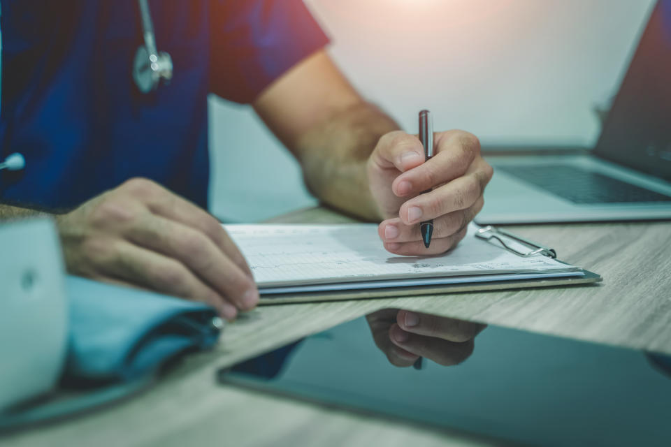 Doctor making notes in medical journal using laptop computer sitting at desk in clinic.