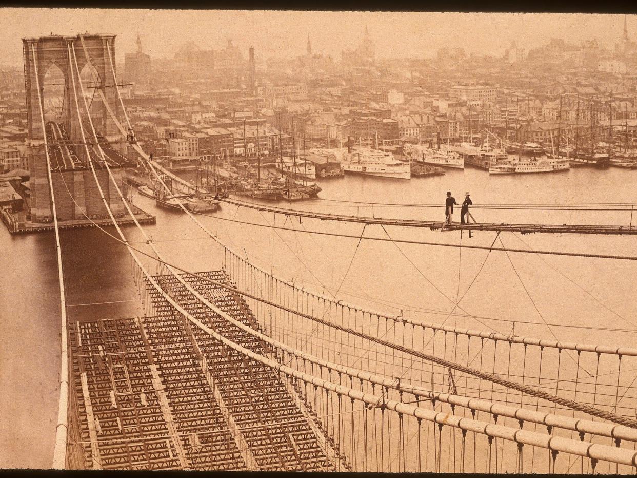 Two men standing on a high catwalk, surveying the construction of the Brooklyn Bridge