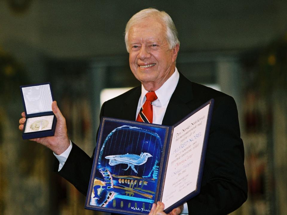 jimmy carter accepts the nobel peace prize in 2002, he holds a small box with the medal inside in his right hand, in his left hand, he holds a certificate for the prize that includes a blue, white and gold drawing, carter is smiling and wearing a black suit jacket, white collard shirt and red tie with a black and white stripe