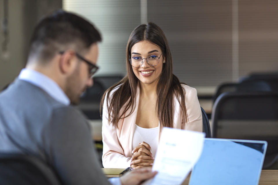 Business, career and placement concept - young woman smiling and holding resume while sitting in front of mentor or managers during job interview. Smiling female client talking to male manager