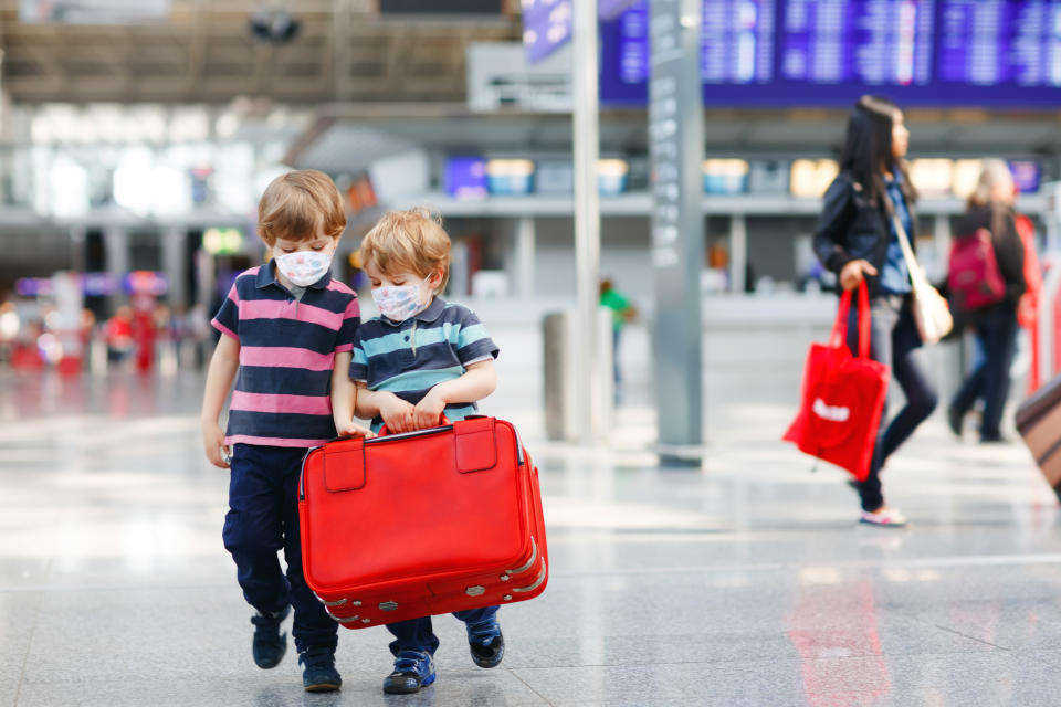 Two little sibling boys wear medical mask and going on vacations trip with suitcase at airport, indoors. Happy twins brothers walking to check-in or boarding for flight during corona virus pandemic.