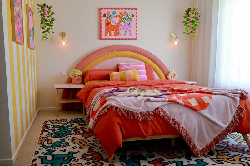 Bedroom with bed with orange and yellow curved fabric headboard, keith haring rug, and yellow and white striped accent wall