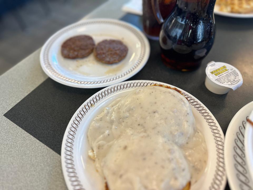 A plate of two large biscuits covered in white gravy with a white plate holding two sausage patties in the background.