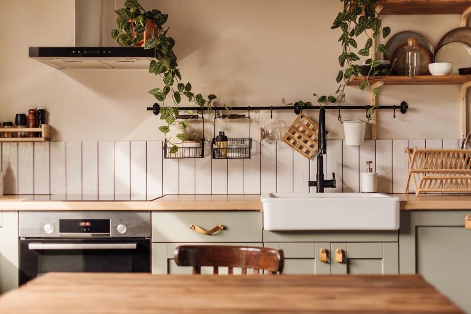 empty kitchen island with marble surface in foreground, green vintage countertop with drawers and pendant lights hanging above, lots of flowers in jars, blurred background