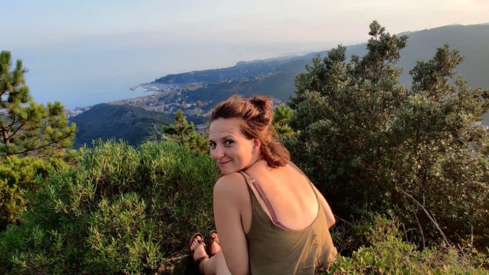 Megan Morgan sentada em uma montanha, olhando por cima do ombro para a câmera. Ao longe, é possível ver arbustos e o mar