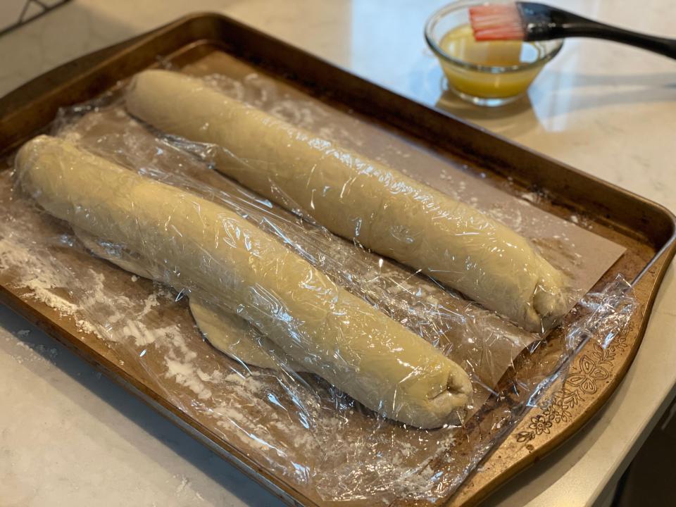 two uncooked baguettes prooving on a cookie sheet covered in plastic