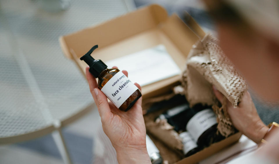 A hand holding a bottle labeled "face cleanser" next to an open cardboard box with packing material and other cosmetic items