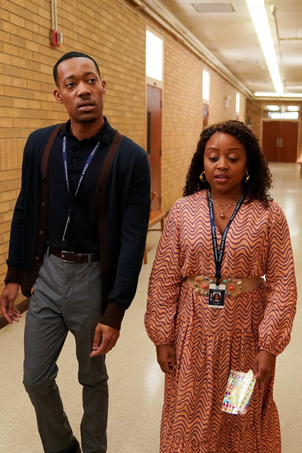 Two individuals walking down a school hallway, both wearing professional attire with ID badges