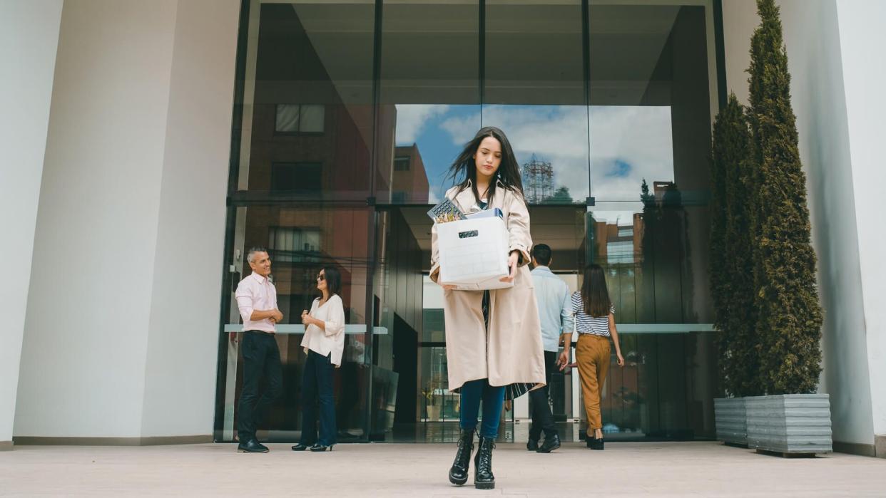 Fired woman leaving the office building with her belongings in a box and looking sad - losing a job concepts.