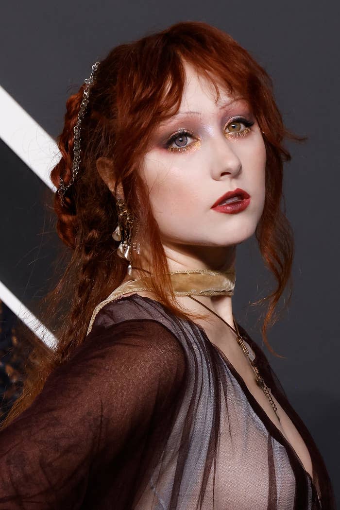 Woman with red hair, smoky eye makeup, and red lipstick wears a sheer, patterned dress and an elaborate updo