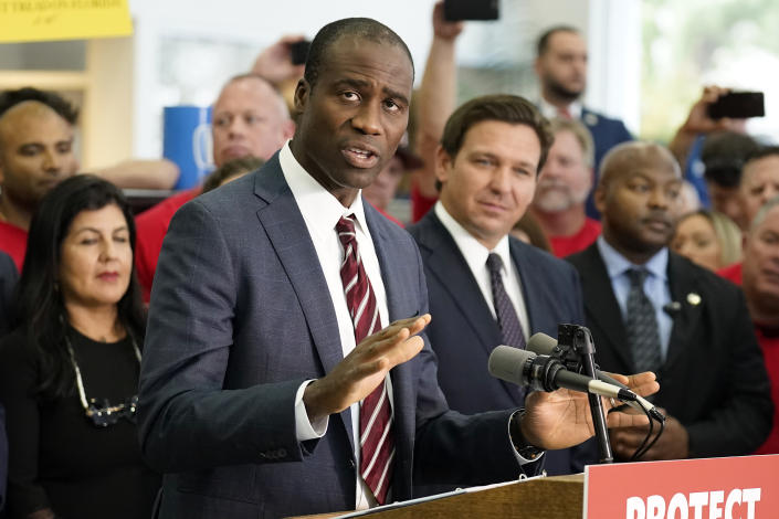 Florida’s surgeon general, Dr. Joseph Ladapo speaking from a podium as Gov. Ron DeSantis looks on.