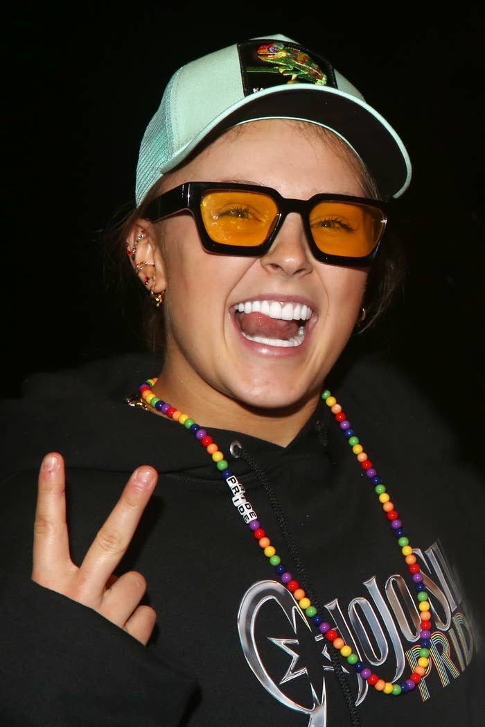 JoJo Siwa wears tinted glasses, a trucker hat, a hoodie, and a beaded rainbow necklace while smiling and flashing a peace sign