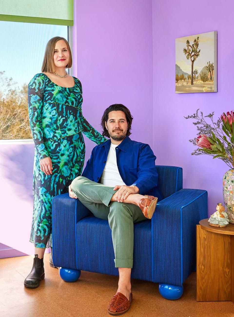 designer in a green patterned dress standing next to husband seated in a dark denim like blue armchair against a purple wall and wood floors