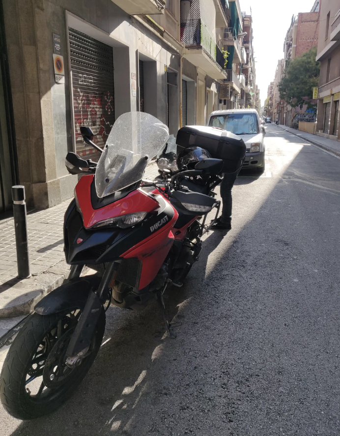 A person stands beside a parked motorcycle labeled "Bicicati" on a quiet city street. The person is facing away from the camera