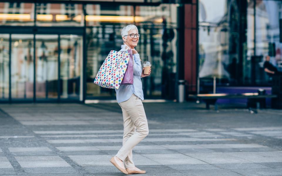 Smiling mature woman holding coffee and walking with shopping bags in the city - VioletaStoimenova