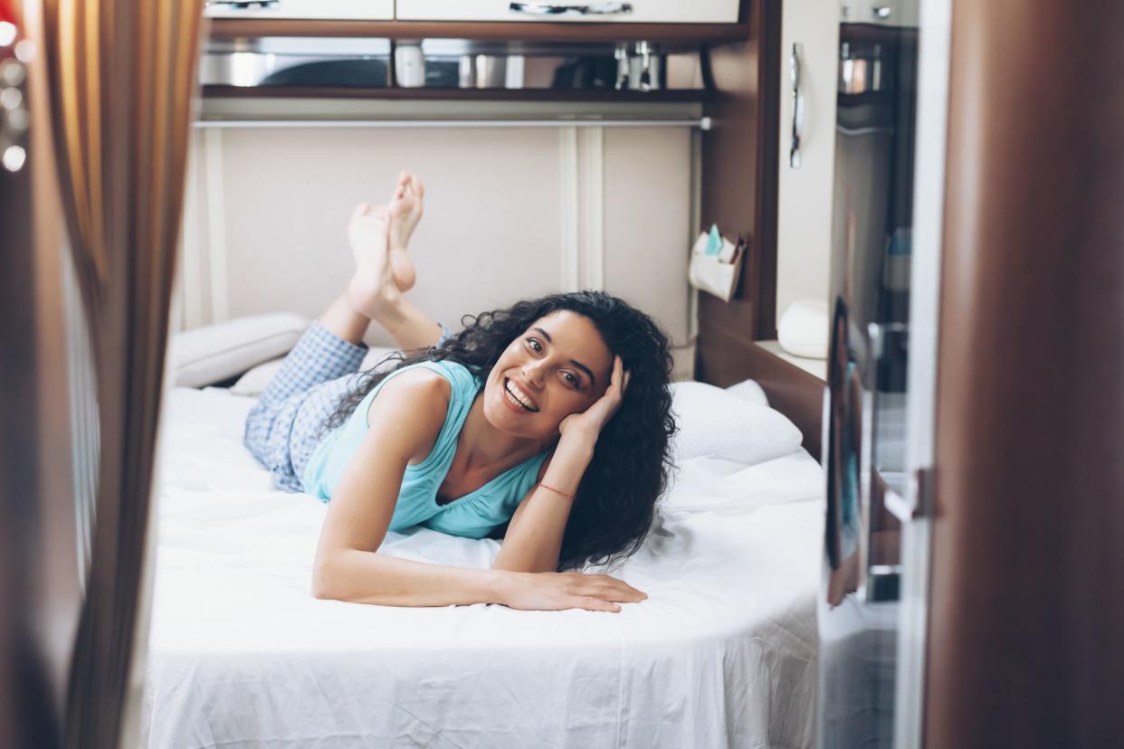 Young woman lying on front on bed in camper and resting. With long black hair and casual clothes. Looking at camera.