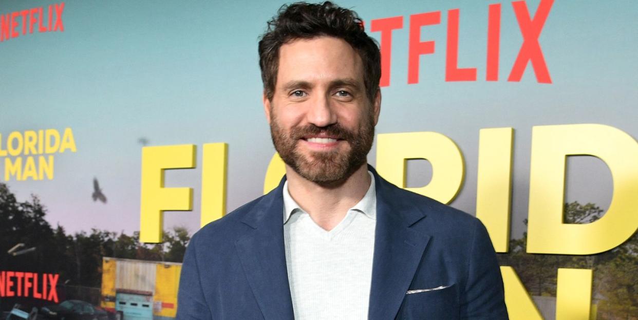 edgar ramirez, a man stands looking at the camera smiling, he has brown hair and a beard, he wears a white top with blue suit