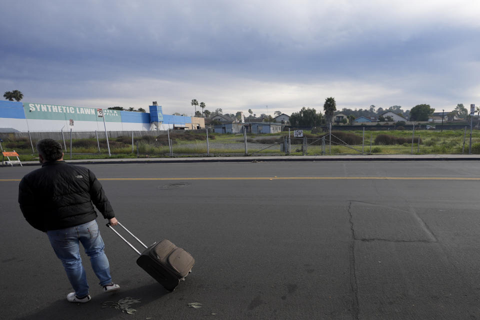 A migrant rolls a suitcase as he arrives at a bus stop after leaving a processing facility, Friday, Feb. 23, 2024, in San Diego. Hundreds of migrants were dropped off Friday at a sidewalk bus stop amid office parks in San Diego with notices to appear in immigration court after local government funding for a reception center ran out of money sooner than expected. (AP Photo/Gregory Bull)