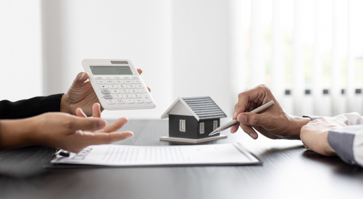 An image of two people with a housing contract, hands holding a pen, hands holding a calculator with a house in the background