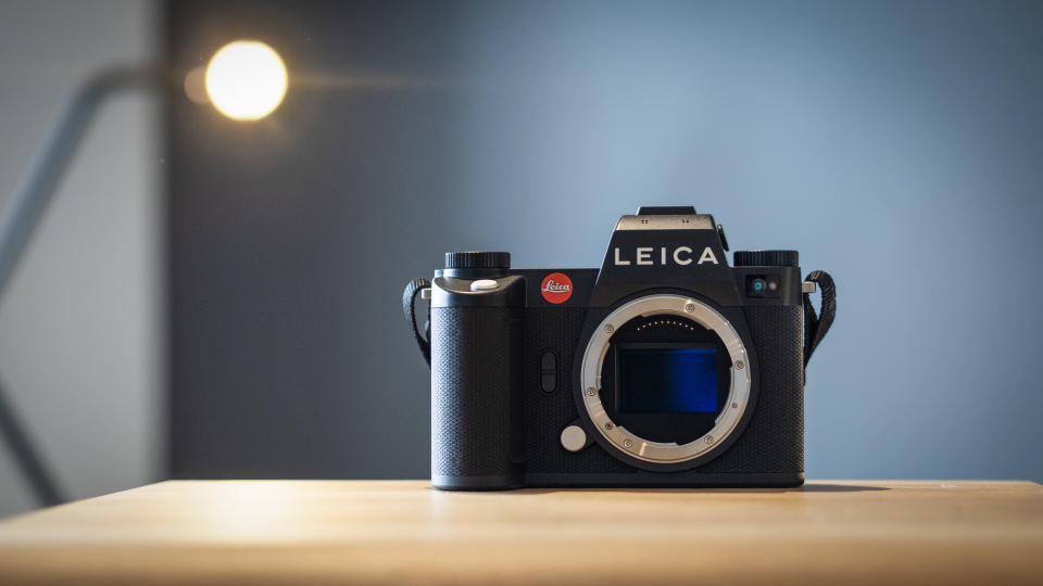 A Leica SL3 camera, with the sensor showing, sitting on a wooden surface in front of a blue background with a small spotlight