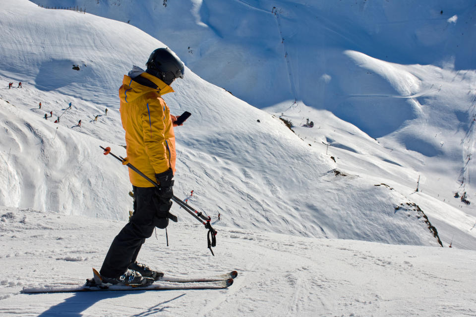 Skier using smart phone on top snow mountain practising ski sport. Skier pauses to check smart phone for reading a phone information. Horizontal photgraphy