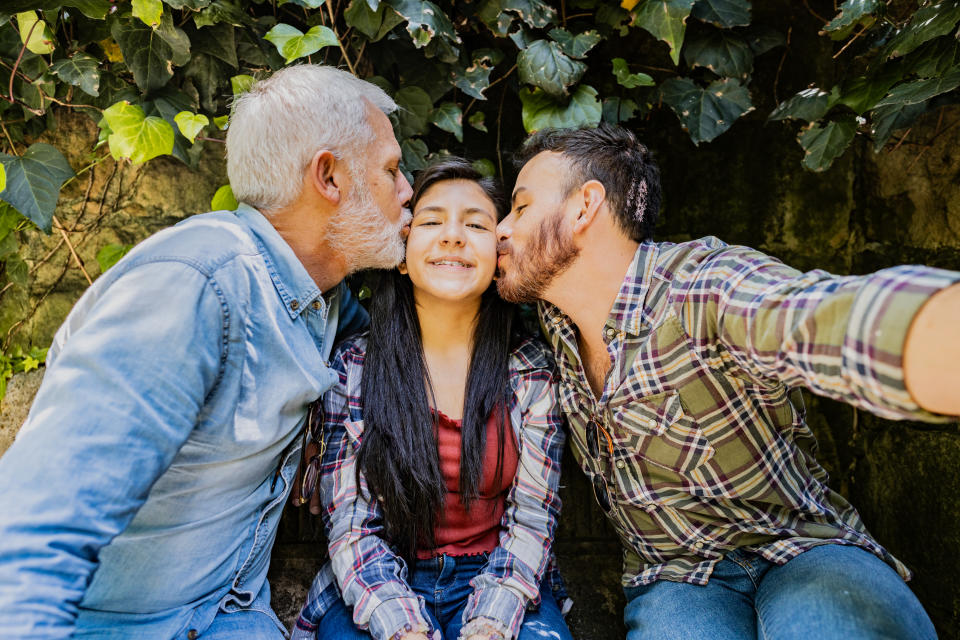 Two men kiss a smiling young girl on her cheeks as they sit outdoors