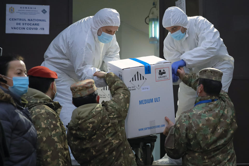 Men wearing full protective suits against coronavirus, take a box containing COVID-19 vaccines, from military personnel at the National Center for Storage of the COVID-19 Vaccine, a military run facility, in Bucharest, Romania, Saturday, Dec. 26, 2020. Romanian authorities will start COVID-19 vaccinations on Dec. 27, 2020. (AP Photo/Vadim Ghirda)