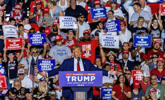 Donald Trump at podium with supporters holding signs behind him