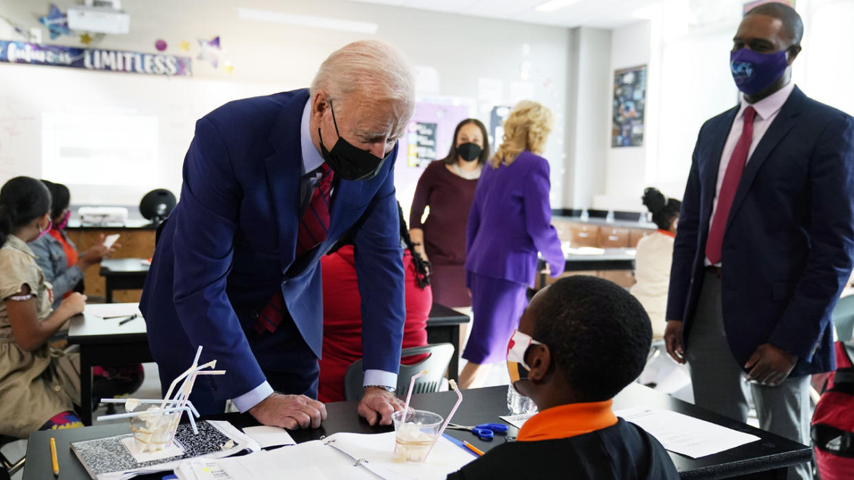 President Biden in a classroom, leans over a student's desk in conversation, while Jill Biden and other adults are seen behind him, all wearing face masks.