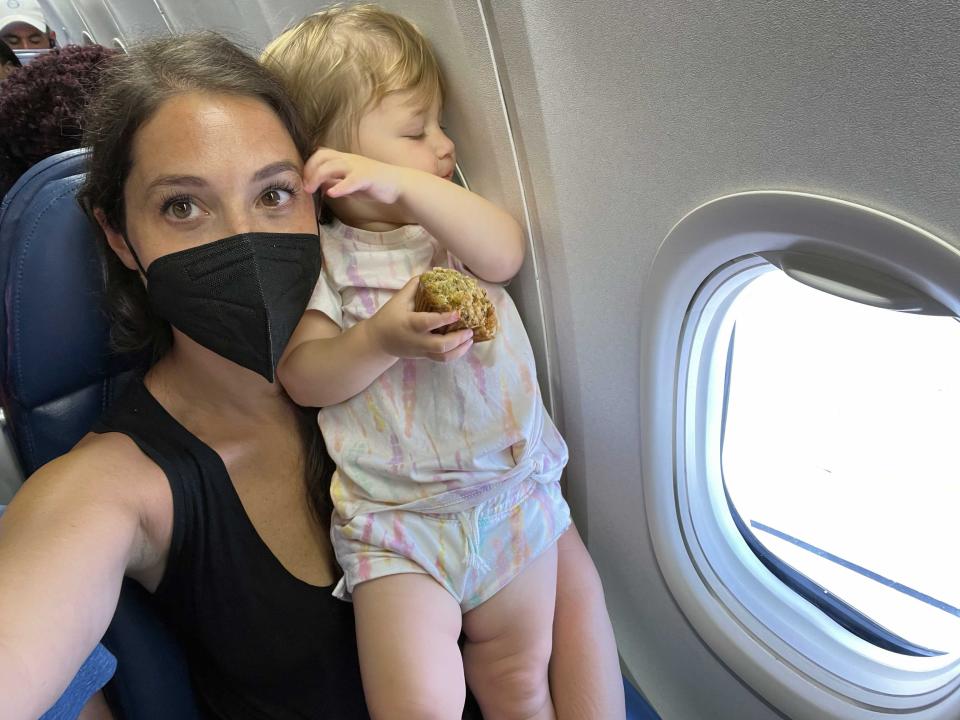 A woman taking a selfie on an airplane holding her toddler next to the window who is holding a muffin.