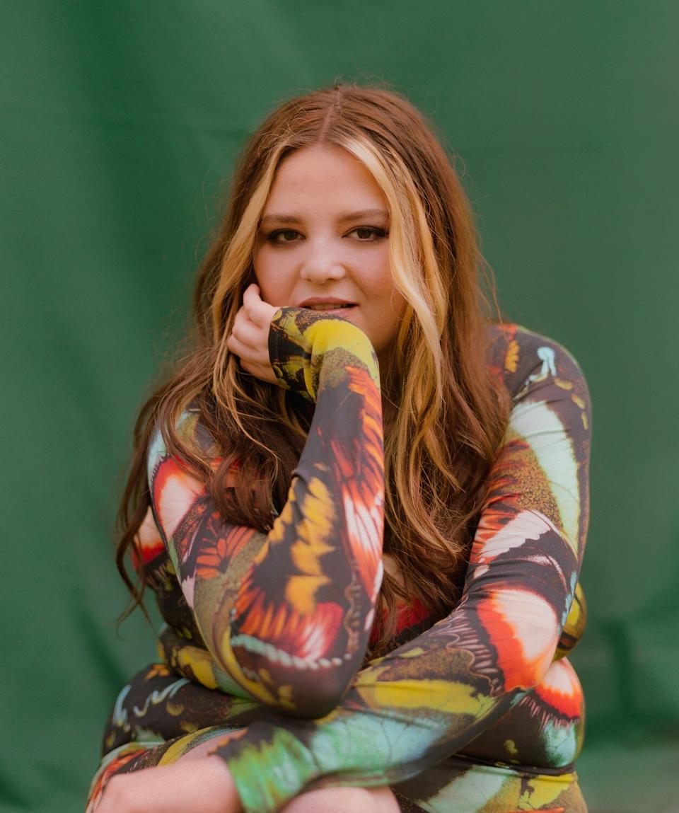 A woman in a colorful shirt poses for a portrait in front of a green fabric background.