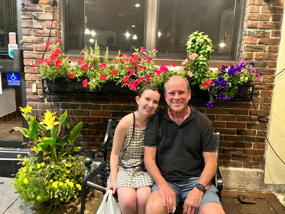  Image of Natalie Daniels on the left with her father on the right, sitting on a black metal bench outside a business, which has a flower box outside its window with red and purple flowers. Natalie has her dark brown hair pulled back and wears a black and white checkered dress. She holds a white plastic bag in her right hand and sits close to her dad. Her dad's shoulder has grey hair and a mostly bald head with light blue eyes. He wears a black polo shirt. 